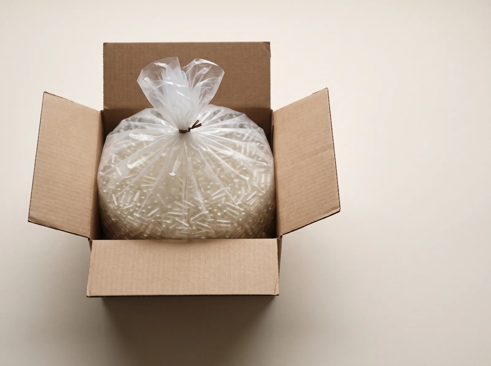 An open cardboard carton containing a tied clear inner bag of empty capsules.