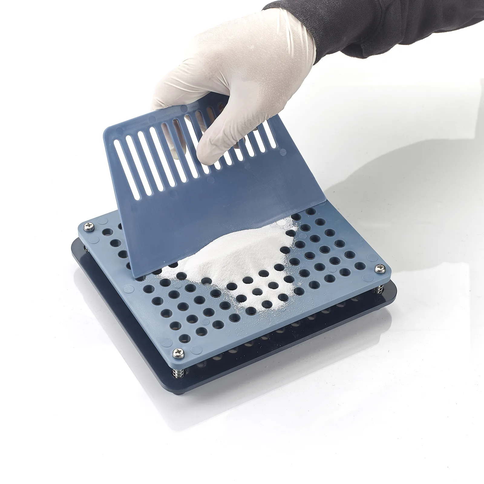 A gloved hand lifting a slotted component above a blue manual capsule-filling plate.